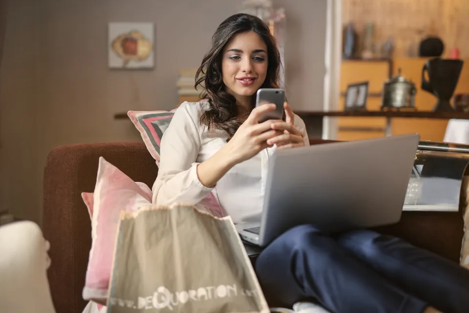 Koltukta oturmuş, telefon ve dizüstü bilgisayarla online alışveriş yapan bir kadın